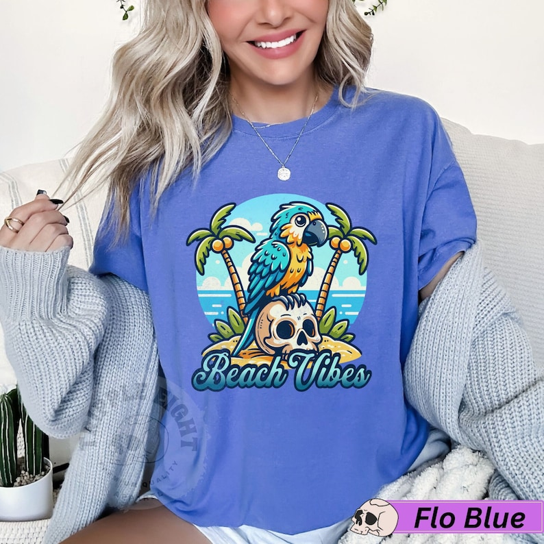 May include: A blue and yellow parrot sits on a skull with palm trees in the background. The text "Beach Vibes" is written below the parrot. The shirt is a light blue color.