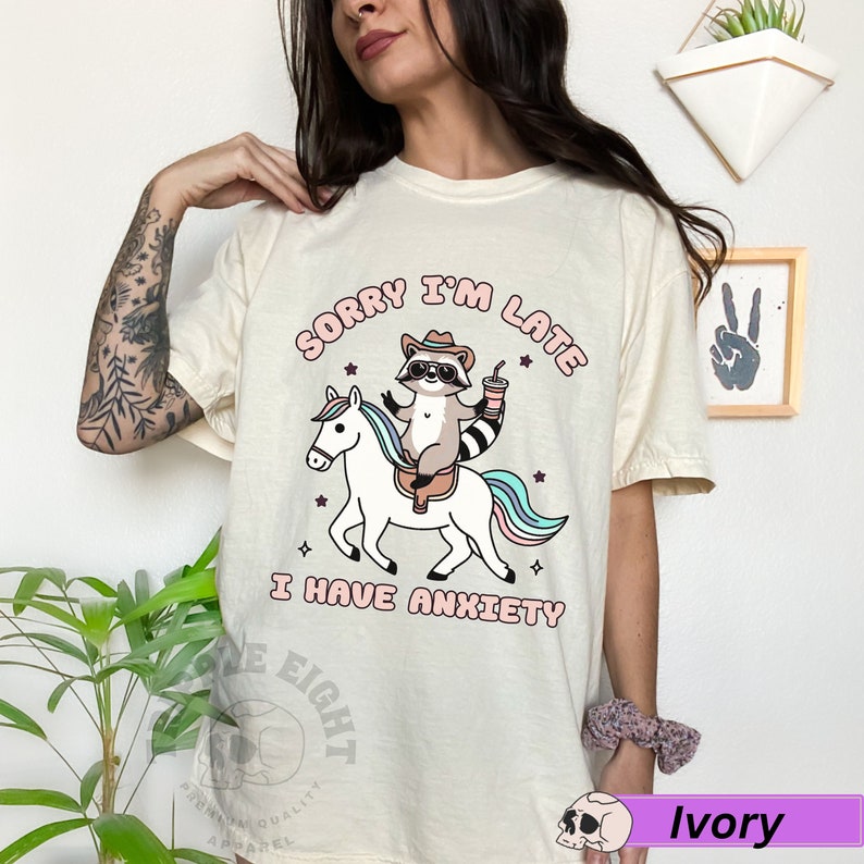 May include: A white t-shirt with a cartoon raccoon riding a unicorn. The text on the shirt reads "Sorry I'm Late I Have Anxiety".