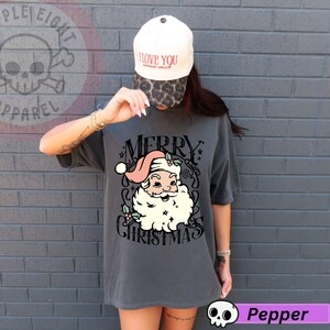 May include: A charcoal gray t-shirt featuring a vintage-style Santa Claus graphic with the words "MERRY CHRISTMAS". The Santa Claus illustration has a pink hat and a white beard. The person is wearing a white baseball cap with the words "I LOVE YOU" on it.