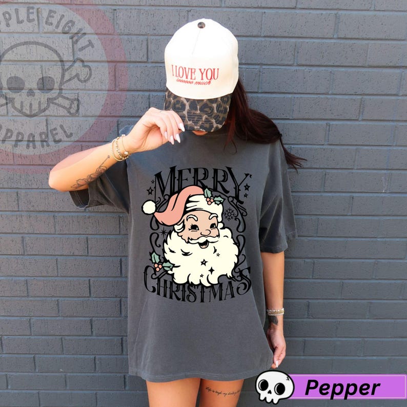 May include: A charcoal gray t-shirt featuring a vintage-style Santa Claus graphic with the words "MERRY CHRISTMAS". The Santa Claus illustration has a pink hat and a white beard. The person is wearing a white baseball cap with the words "I LOVE YOU" on it.