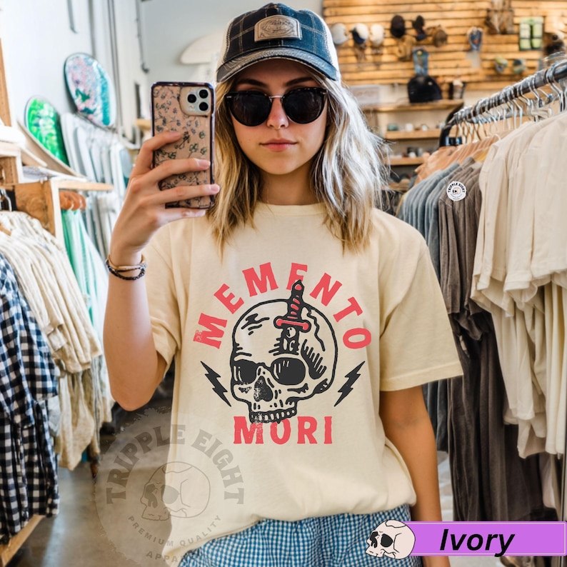 Puede incluir: Una mujer lleva una camiseta beige claro con un gr&aacute;fico de calavera y el texto "Memento Mori" en rojo. La calavera tiene una daga que la atraviesa. La camiseta es de estilo unisex.