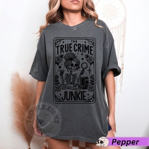 Puede incluir: Una camiseta gris oscuro con un dise&ntilde;o gr&aacute;fico negro de una mujer esqueleto sosteniendo una copa de vino y una lupa. El gr&aacute;fico est&aacute; rodeado de rosas y estrellas. El texto "THE TRUE CRIME JUNKIE" est&aacute; impreso en el gr&aacute;fico.
