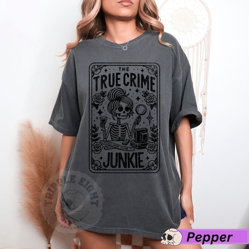 Puede incluir: Una camiseta gris oscuro con un dise&ntilde;o gr&aacute;fico negro de una mujer esqueleto sosteniendo una copa de vino y una lupa. El gr&aacute;fico est&aacute; rodeado de rosas y estrellas. El texto "THE TRUE CRIME JUNKIE" est&aacute; impreso en el gr&aacute;fico.