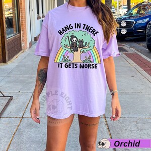 May include: A purple t-shirt with the text "Hang in there it gets worse" printed on it. The graphic features a cartoon grim reaper sitting on a tree with a cat. A rainbow is behind the tree.