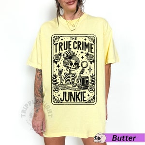 Puede incluir: Una camiseta amarilla con un gr&aacute;fico negro de un esqueleto sosteniendo una copa de vino y una lupa. El gr&aacute;fico est&aacute; en un marco rectangular con el texto "THE TRUE CRIME JUNKIE" en la parte superior.