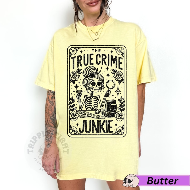 Puede incluir: Una camiseta amarilla con un gr&aacute;fico negro de un esqueleto sosteniendo una copa de vino y una lupa. El gr&aacute;fico est&aacute; en un marco rectangular con el texto "THE TRUE CRIME JUNKIE" en la parte superior.