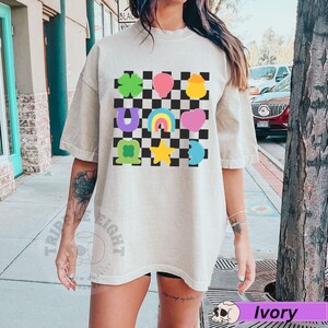 May include: Ivory-colored t-shirt featuring a checkered pattern with colorful cartoon graphics. The graphics include a four-leaf clover, a pink heart, a rainbow, and a yellow star. The shirt has short sleeves and a relaxed fit.