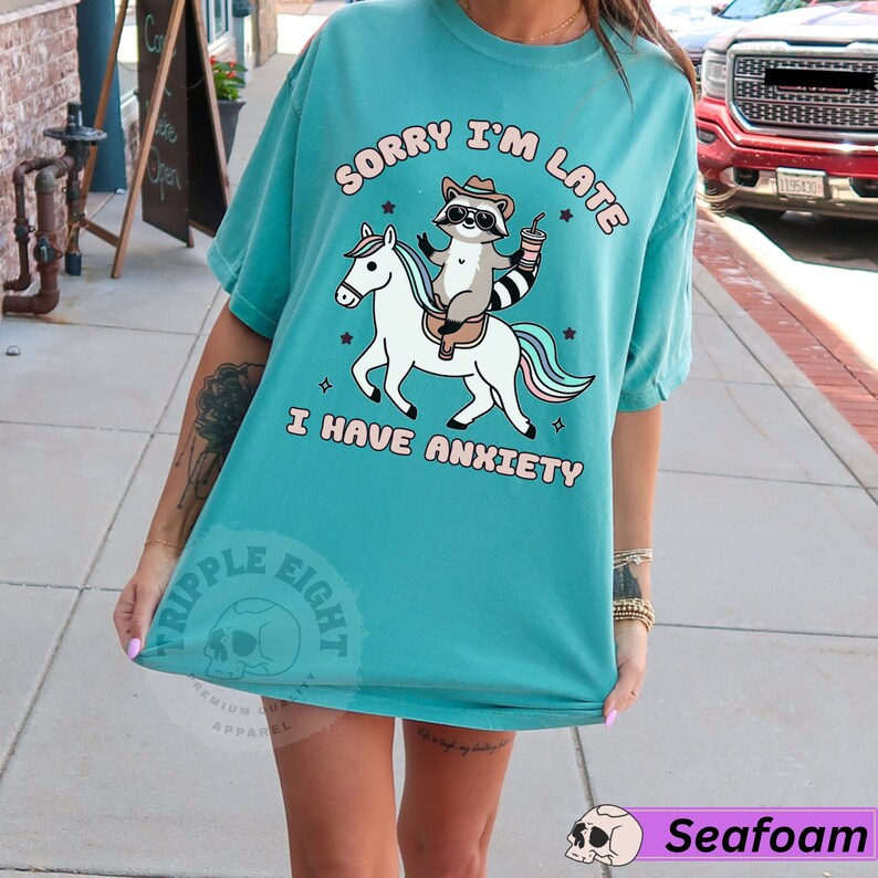 May include: A turquoise blue t-shirt with a graphic of a raccoon riding a unicorn. The text "Sorry I'm Late I Have Anxiety" is printed on the shirt.