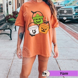 May include: Orange t-shirt featuring a design of three Halloween buckets with jack-o-lantern faces. The buckets are green, white and orange.
