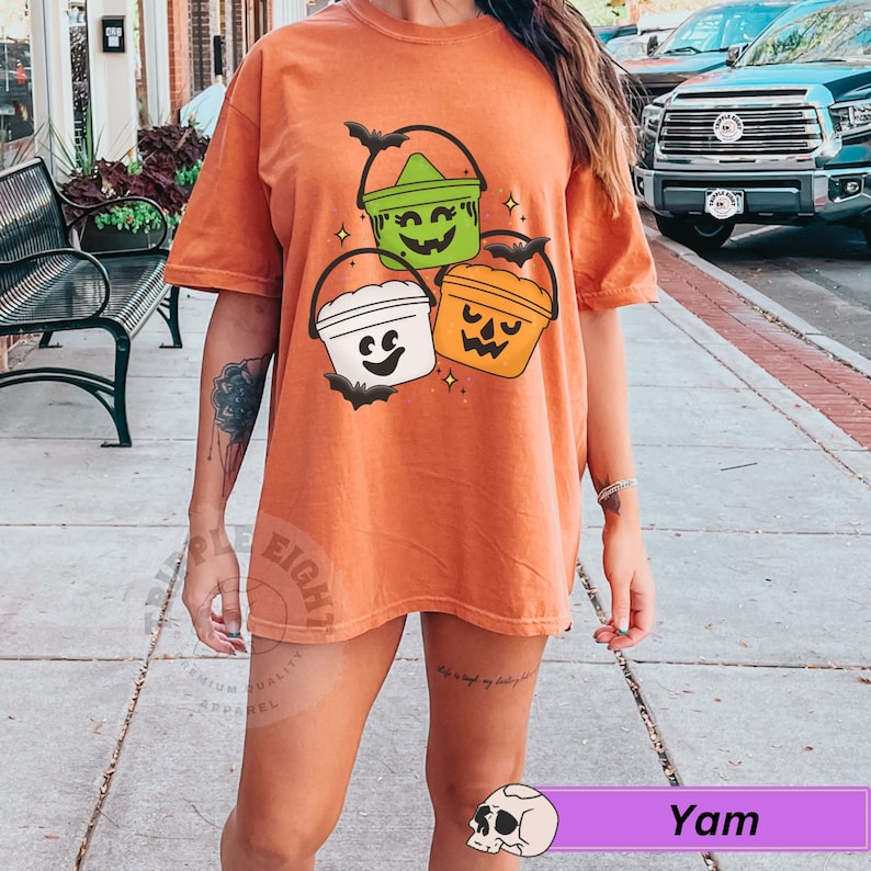 May include: Orange t-shirt featuring a design of three Halloween buckets with jack-o-lantern faces. The buckets are green, white and orange.