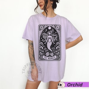 May include: A lavender t-shirt featuring a black and white illustration of a mermaid in a vintage tarot card style. The text "The Mermaid" is printed below the image.
