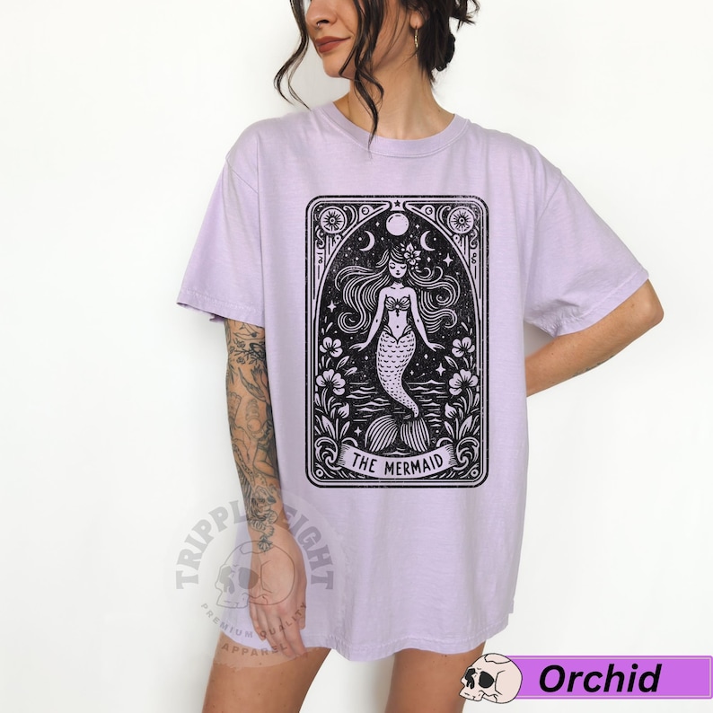 May include: A lavender t-shirt featuring a black and white illustration of a mermaid in a vintage tarot card style. The text "The Mermaid" is printed below the image.
