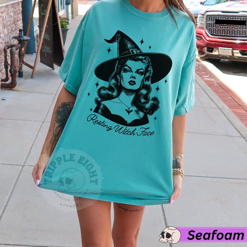 May include: A turquoise blue t-shirt with a black and white illustration of a witch with the text "Resting Witch Face".