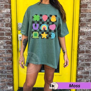 May include: Moss green t-shirt with a colorful graphic of lucky charms on a black and white checkered background. The graphic includes a four-leaf clover, horseshoe, rainbow, heart, and star. The shirt is a comfortable, oversized fit.