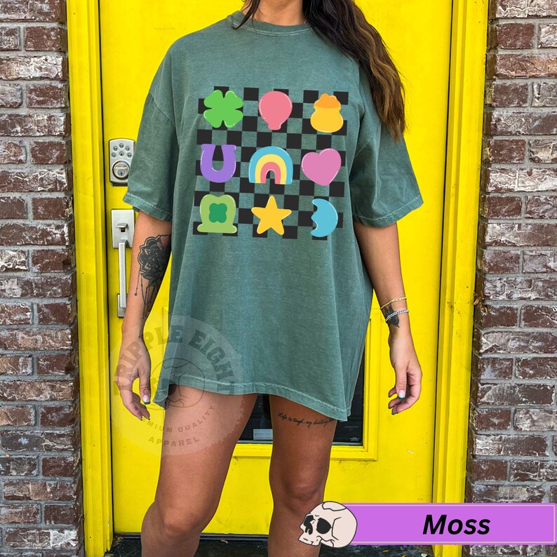 May include: Moss green t-shirt with a colorful graphic of lucky charms on a black and white checkered background. The graphic includes a four-leaf clover, horseshoe, rainbow, heart, and star. The shirt is a comfortable, oversized fit.