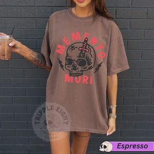 Puede incluir: Una camiseta marr&oacute;n con un gr&aacute;fico de calavera y el texto "Memento Mori" en rojo. La calavera tiene una daga atravesando su cabeza. La camiseta es de la marca "Triple Eight".