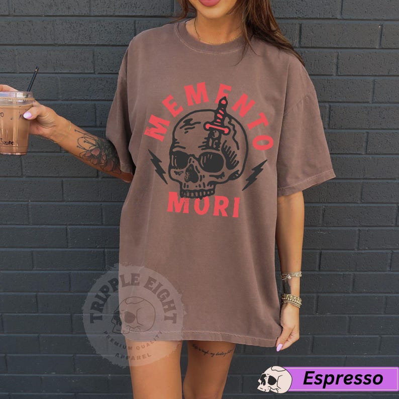 Puede incluir: Una camiseta marr&oacute;n con un gr&aacute;fico de calavera y el texto "Memento Mori" en rojo. La calavera tiene una daga atravesando su cabeza. La camiseta es de la marca "Triple Eight".