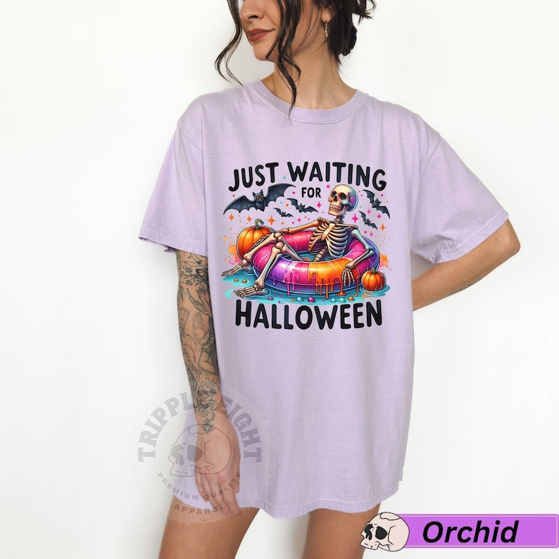 May include: A lavender t-shirt with the text "Just Waiting for Halloween" and a skeleton wearing a colorful inflatable ring. The skeleton is surrounded by bats, pumpkins, and candy.