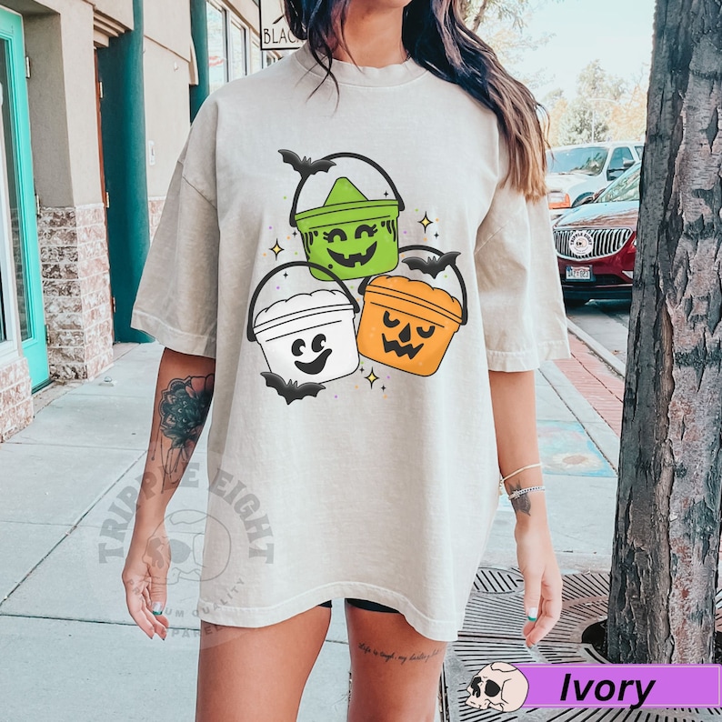 May include: A light beige t-shirt featuring a graphic of three Halloween buckets with faces. The buckets are green, white, and orange. The graphic also includes bats and stars.