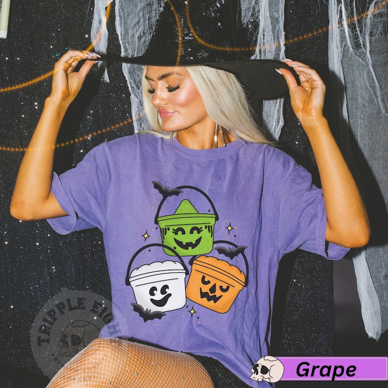 May include: Purple t-shirt featuring a graphic of three Halloween buckets with jack-o-lantern faces. The buckets are green, white, and orange. The shirt has the text "Triple Eight" on the left side.