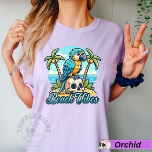 May include: A purple t-shirt with a cartoon parrot perched on a skull with palm trees and the text "Beach Vibes". The shirt has a logo on the bottom left corner that says "Triple Eight Premium Quality Apparel".