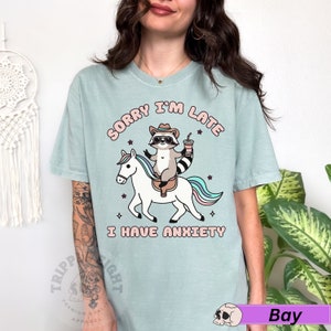 May include: A light blue t-shirt with a graphic of a raccoon wearing a cowboy hat and riding a unicorn. The text on the shirt reads "Sorry I'm Late I Have Anxiety".