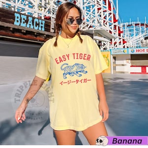 May include: Pale yellow t-shirt featuring the text "EASY TIGER" above a blue and white tiger illustration. Japanese text is below the tiger. The shirt is from the "Banana" collection.