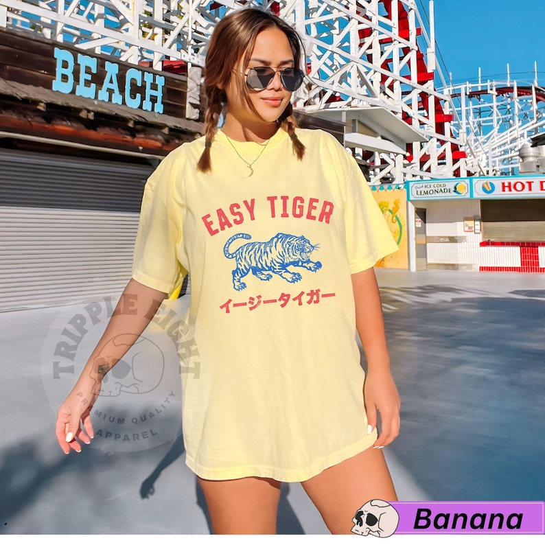 May include: Pale yellow t-shirt featuring the text "EASY TIGER" above a blue and white tiger illustration. Japanese text is below the tiger. The shirt is from the "Banana" collection.