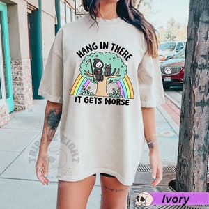 May include: A light beige t-shirt with a graphic of a skeleton sitting in a tree with a cat. The text on the shirt reads "Hang in there it gets worse".