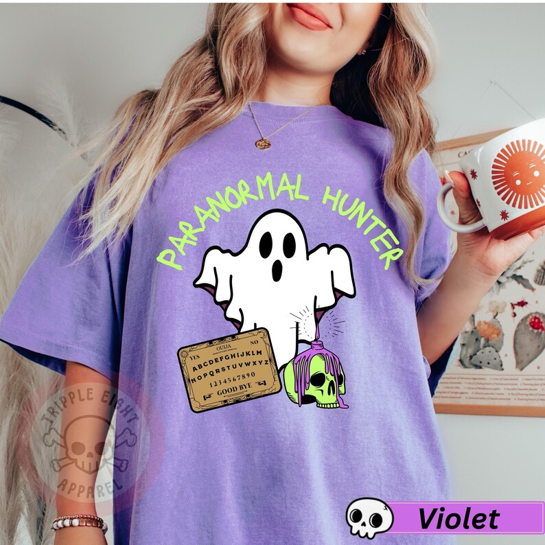 May include: Violet-colored t-shirt with a graphic of a white ghost, a Ouija board, and a green skull with dripping purple slime. The text "PARANORMAL HUNTER" is in green above the ghost. The shirt also has the text "TRIPPLE EIGHT APPAREL".