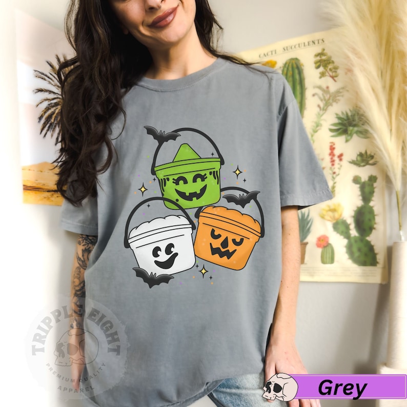 May include: A grey t-shirt featuring a design of three Halloween buckets with different coloured lids and faces. The buckets are green, white, and orange. The shirt has the text "Tripple Eight" in the bottom left corner.