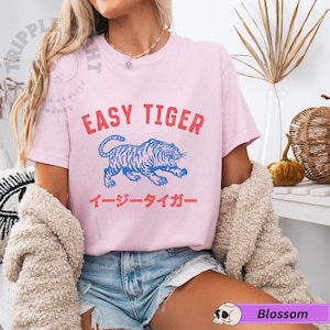 May include: A pale pink t-shirt featuring the words "EASY TIGER" in red, above a blue tiger illustration. Below the tiger is Japanese text in red. The shirt has short sleeves.