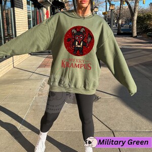 May include: A green sweatshirt with a red circle graphic of a cartoon Krampus holding a torch. The text "Merry Krampus" is printed in red below the graphic.