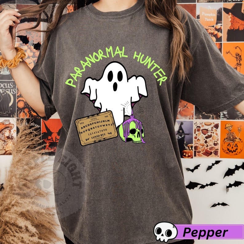 May include: Dark gray t-shirt with a graphic featuring a white ghost holding a green skull and a Ouija board. The words "PARANORMAL HUNTER" are printed above the ghost in green. A fun Halloween-themed design.