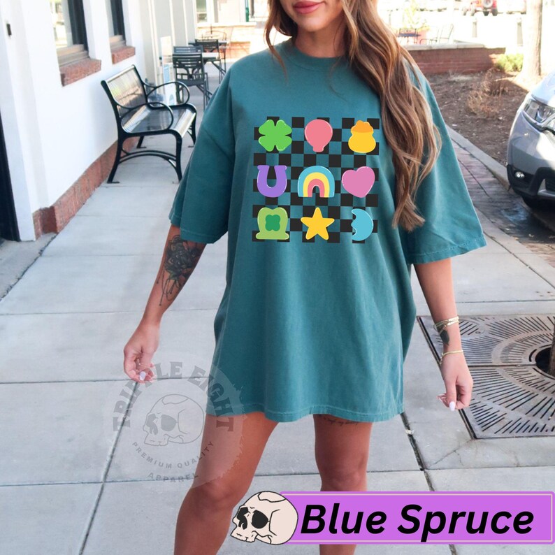 May include: Blue Spruce oversized t-shirt featuring a colorful graphic design with a checkerboard pattern and various symbols, including a four-leaf clover, balloons, and a rainbow. The shirt has short sleeves and a relaxed fit.
