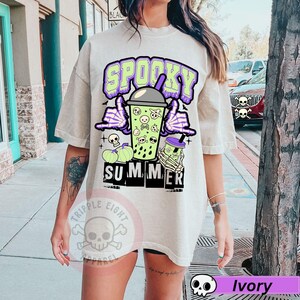 Puede incluir: Camiseta color marfil con un dise&ntilde;o de verano espeluznante. El dise&ntilde;o presenta una bebida verde con calaveras, manos de esqueleto y las palabras "Spooky Summer" en morado y verde. Incluye calabazas y murci&eacute;lagos.