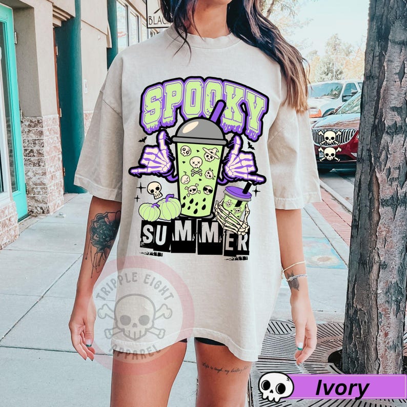 Puede incluir: Camiseta color marfil con un dise&ntilde;o de verano espeluznante. El dise&ntilde;o presenta una bebida verde con calaveras, manos de esqueleto y las palabras "Spooky Summer" en morado y verde. Incluye calabazas y murci&eacute;lagos.