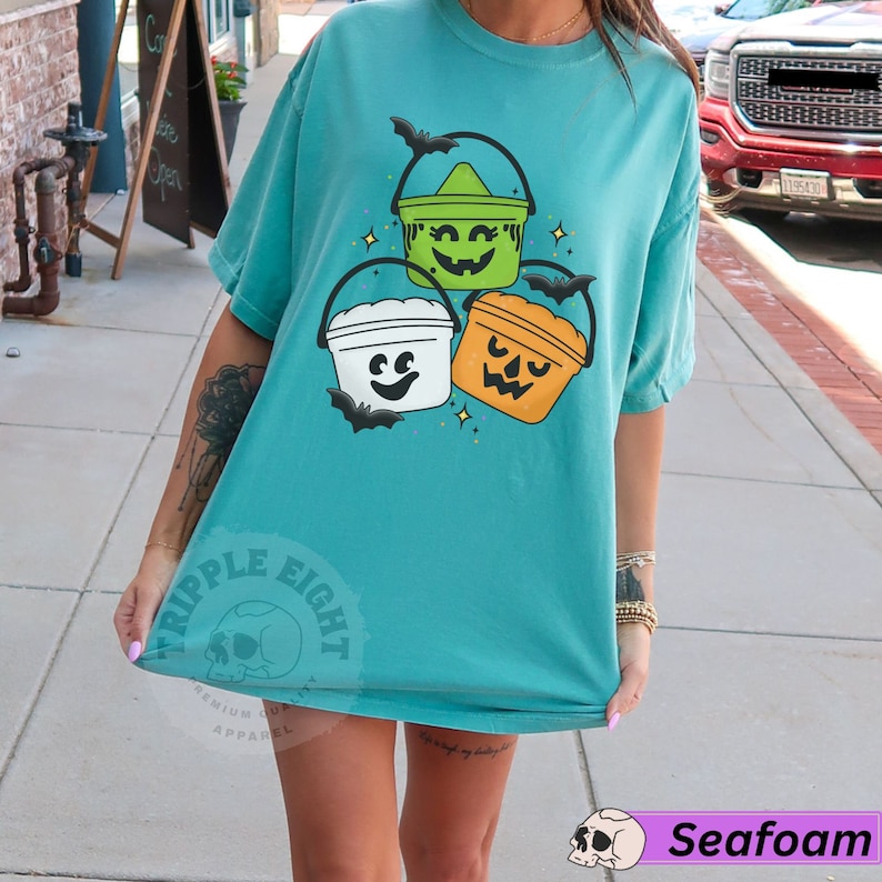 May include: A teal blue t-shirt featuring a graphic of three Halloween buckets with jack-o'-lantern faces. The buckets are stacked on top of each other and have bats flying around them. The text "Ripple Eight" is visible on the bottom left corner of the shirt.