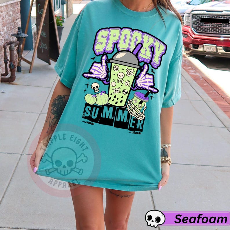 Puede incluir: Camiseta turquesa con un gr&aacute;fico de verano espeluznante. El dise&ntilde;o presenta la palabra "Spooky" en morado, una bebida boba con calaveras, manos de esqueleto y la palabra "Summer". La camiseta tiene un corte holgado.
