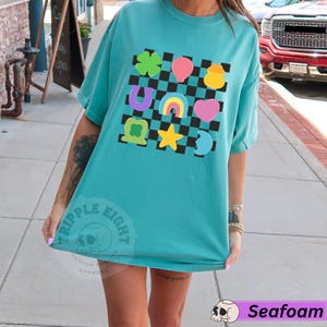 May include: Seafoam green t-shirt with a colorful graphic of lucky charms on a checkered background. The design includes a four-leaf clover, balloon, pot of gold, horseshoe, rainbow, heart, star, and crescent moon. The shirt has the text "TRIPPLE EIGHT" and "Seafoam".