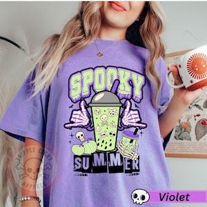 Puede incluir: Camiseta violeta con un dise&ntilde;o de verano espeluznante. El dise&ntilde;o presenta la palabra "Spooky" en verde, una mano de esqueleto, una bebida con calaveras y la palabra "Summer". La camiseta tambi&eacute;n tiene un peque&ntilde;o logotipo de calavera.