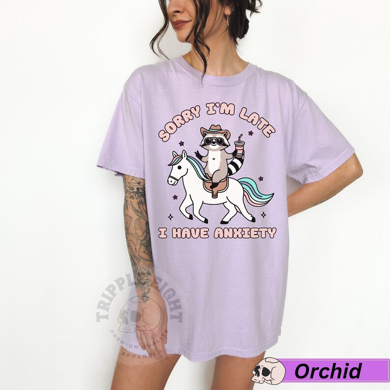 May include: A lavender t-shirt with a graphic of a raccoon riding a unicorn. The text on the shirt reads "Sorry I'm Late I Have Anxiety".