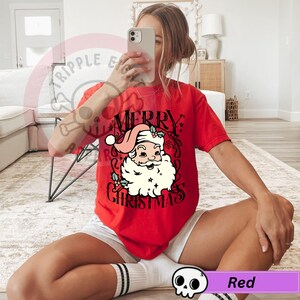 May include: Red t-shirt with a graphic of Santa Claus and the words "Merry Christmas". The Santa Claus graphic features a pink hat, white beard, and holly accents. The shirt is a solid red color.