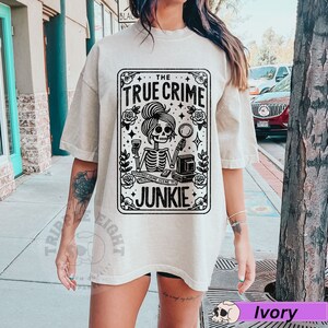 Puede incluir: Una camiseta beige con un gr&aacute;fico negro de una mujer esqueleto sosteniendo una copa de vino y una lupa. El gr&aacute;fico est&aacute; en un marco de tarjeta con el texto "The True Crime Junkie".