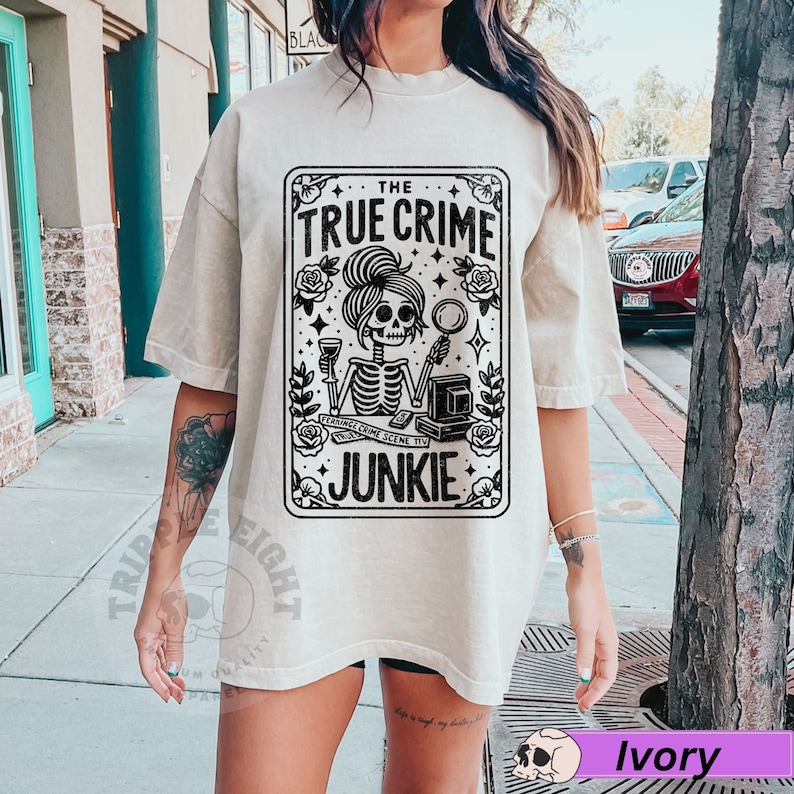 Puede incluir: Una camiseta beige con un gr&aacute;fico negro de una mujer esqueleto sosteniendo una copa de vino y una lupa. El gr&aacute;fico est&aacute; en un marco de tarjeta con el texto "The True Crime Junkie".