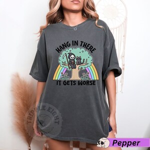 May include: A dark gray t-shirt with a graphic of a grim reaper and a cat sitting on a tree with a rainbow behind them. The text on the shirt reads "Hang in there it gets worse".
