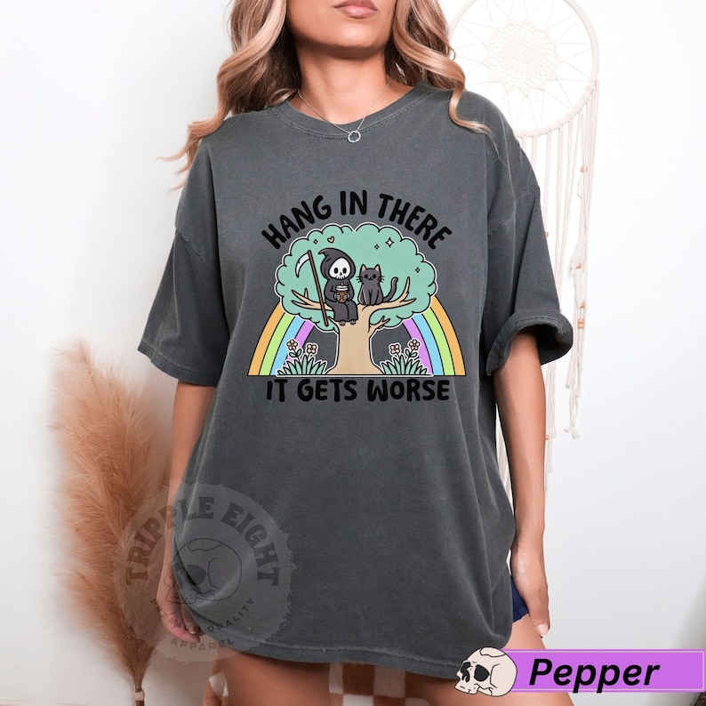 May include: A dark gray t-shirt with a graphic of a grim reaper and a cat sitting on a tree with a rainbow behind them. The text on the shirt reads "Hang in there it gets worse".