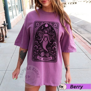 May include: An oversized purple t-shirt featuring a black and white illustration of a mermaid in a vintage tarot card style. The text "The Mermaid" is printed below the image.