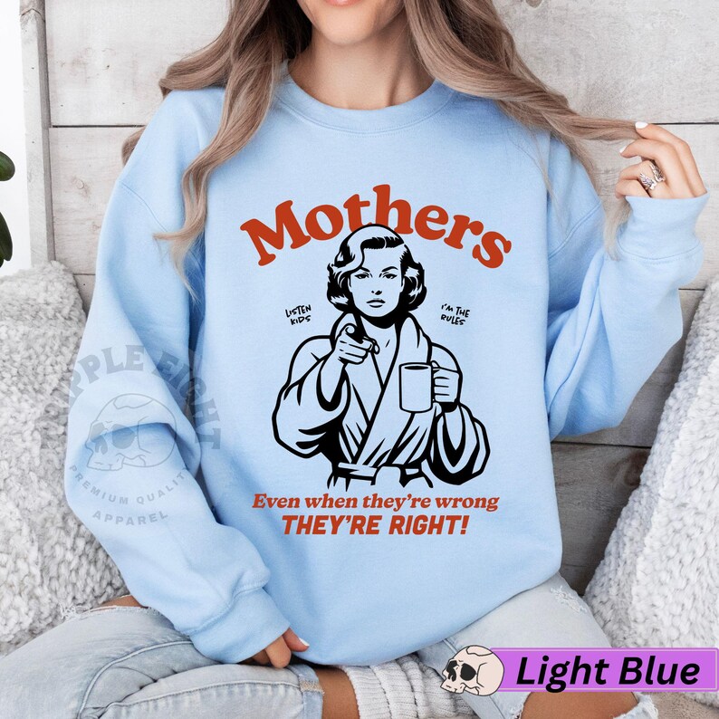 Op de afbeelding: Lichtblauwe trui met een afbeelding van een vrouw in een badjas die een mok vasthoudt. De tekst "Mothers" is rood en de tekst "Listen Kids" en "I'm the Rules" is zwart. De tekst "Even when they're wrong THEY'RE RIGHT!" is rood.