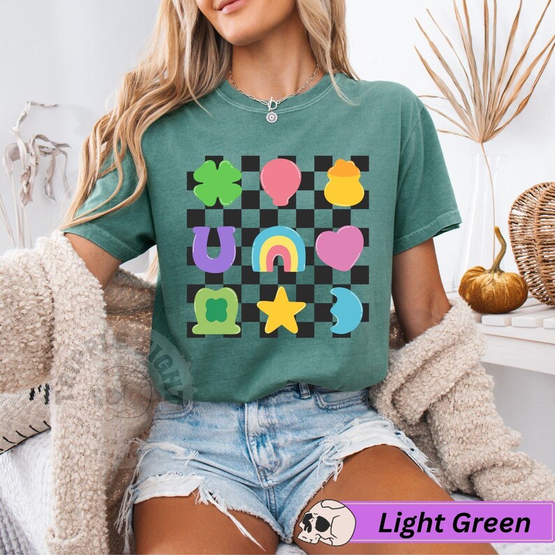 May include: Light green t-shirt with a colorful graphic of lucky charms on a black and white checkered background. The graphic includes a four-leaf clover, horseshoe, rainbow, heart, star, and moon. The text "Light Green" is at the bottom.